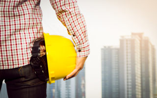Hand's engineer worker holding yellow safety helmet with building on site background.